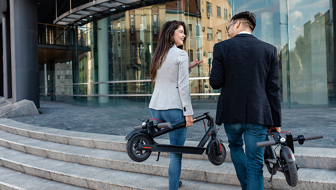 Frau und Mann vor Büroeingang. Sie trägt einen Mini-Roller.